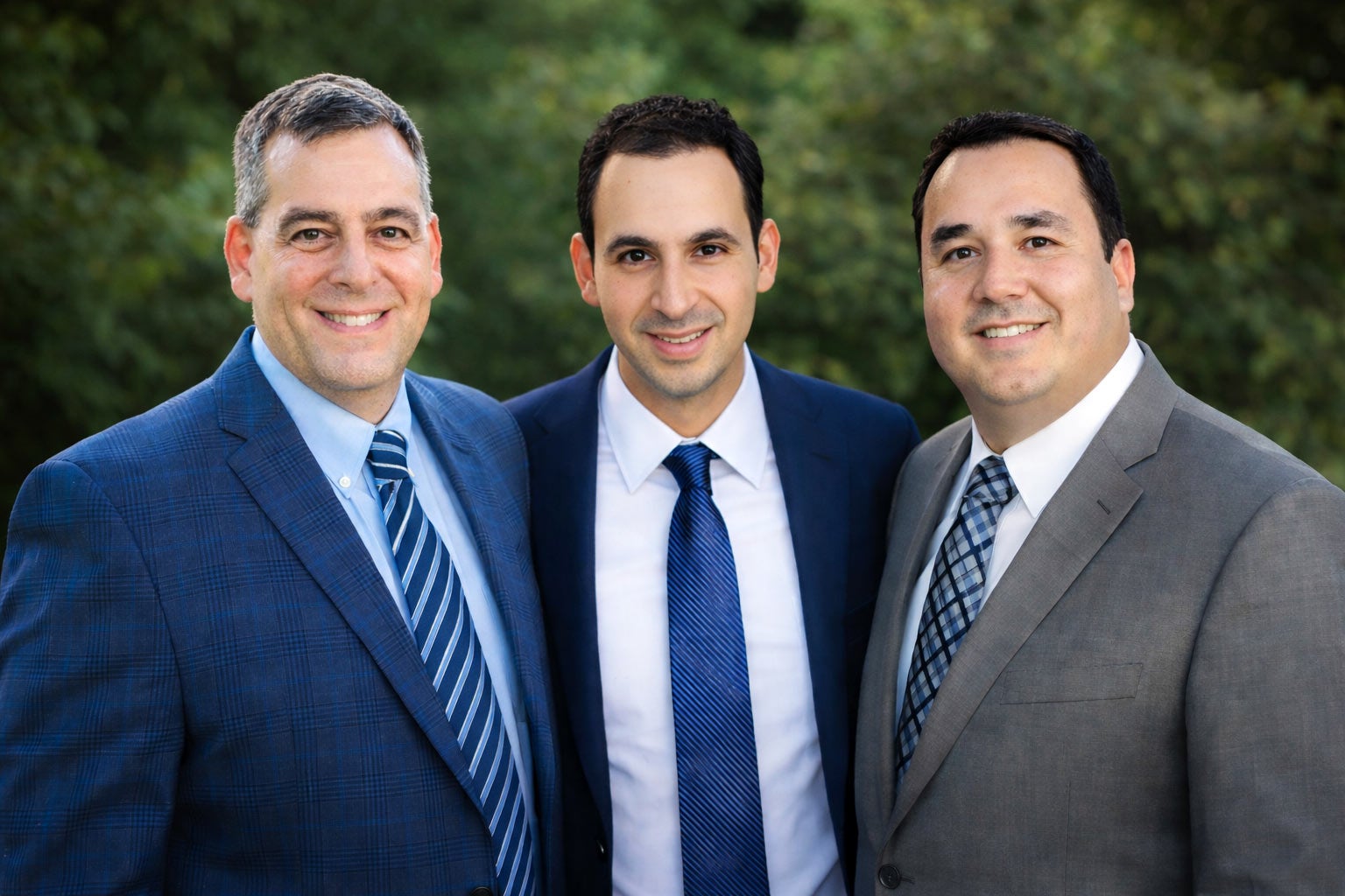 Three men in suits smile and pose closely together outdoors.
