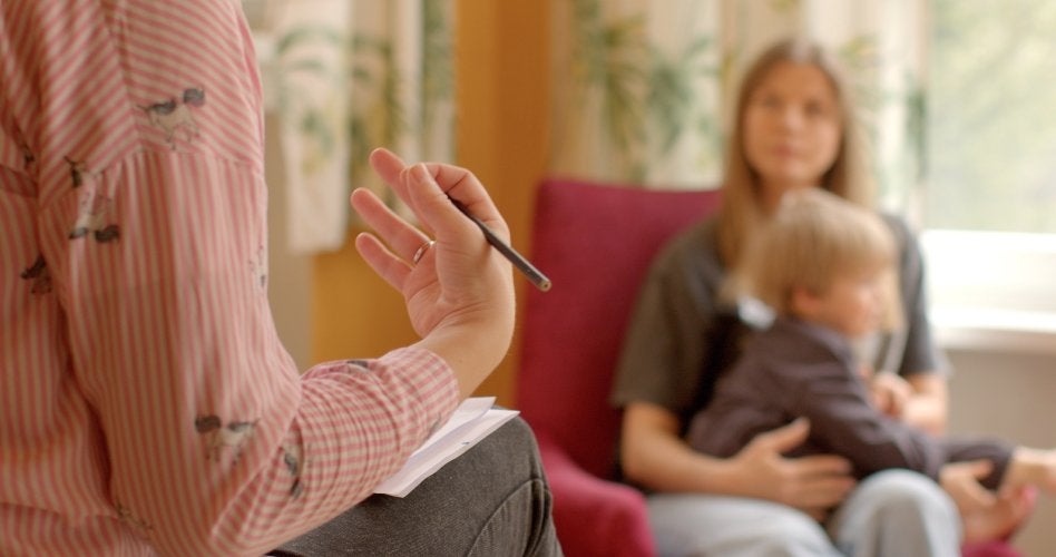 A therapist's hand with a pencil, while a mother with a child watches from the background, out of focus.