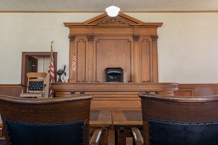 Ornate historic courtroom with wooden judge's bench and two chairs facing it in the foreground.