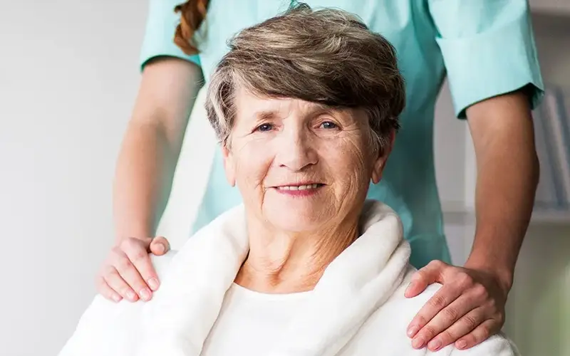 Smiling elderly woman in a white robe with a caregiver's hands on her shoulders.