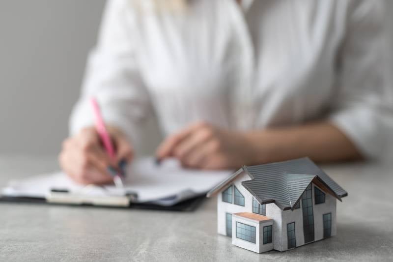 A model house on a table, with a blurred person in a white shirt signing a document in the background.