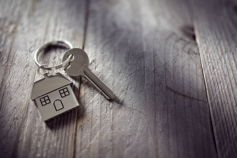 A single house key on a ring with a house-shaped keychain, lying on a weathered gray wooden surface.