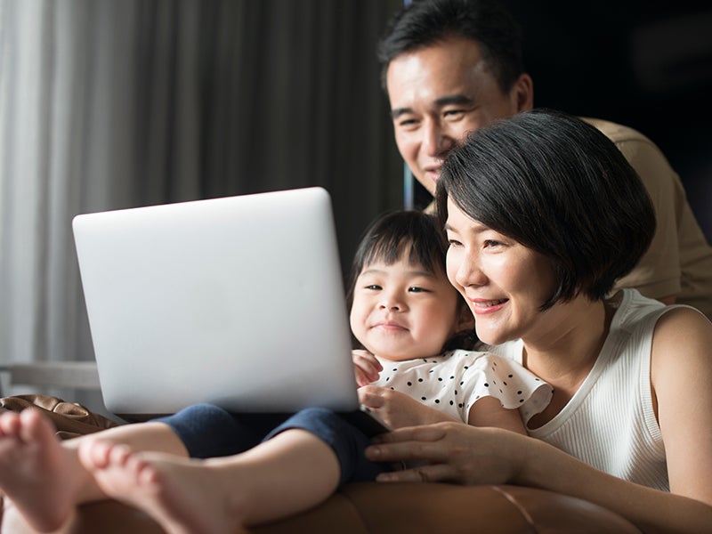 Asian family, including a mother, father, and young child, smiling while looking at a laptop together.