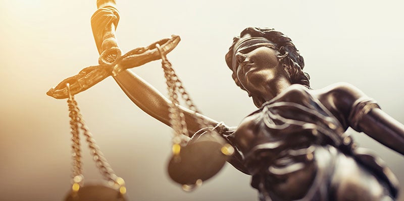 A bronze statue of Lady Justice, blindfolded and holding scales, with a bright light shining in the background.