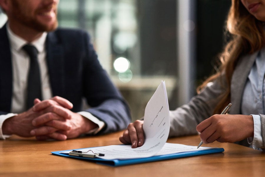 A woman in a gray jacket reviews a document on a blue clipboard while a man in a suit watches.