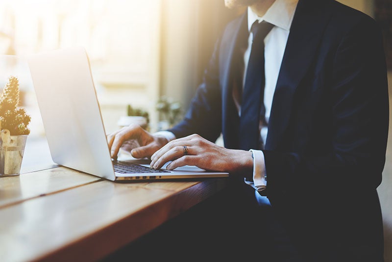 A person in a suit works on a laptop, with hands on the keyboard, on a wooden table.