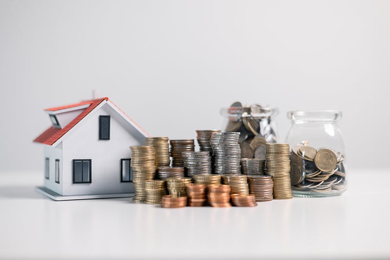 A white model house with a red roof next to stacks of coins and two jars of coins.