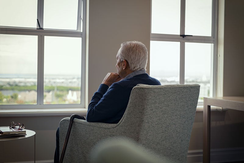 Elderly man with gray hair, seen from behind, sitting in a chair, looking out a window at a distant landscape.