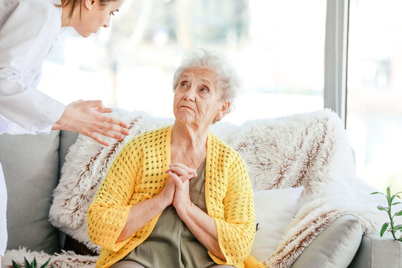 A young female caregiver offers comfort as an elderly woman, sitting on a sofa, looks up anxiously.