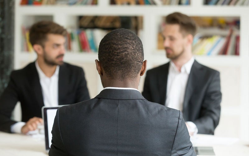 Back of an African-American man in a suit facing two Caucasian men in suits during a job interview.