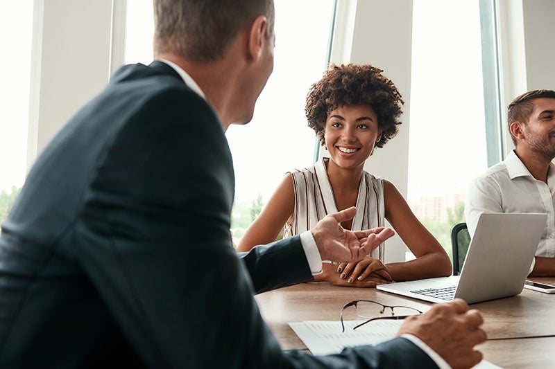 A woman with curly hair smiles while looking at a man in a suit speaking during a meeting.