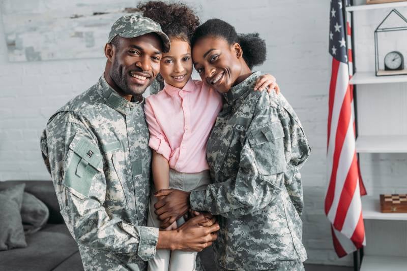 Smiling Black military parents in fatigues with their daughter pose indoors beside an American flag.
