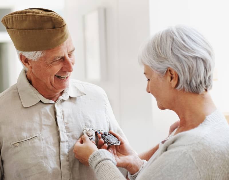 A smiling male veteran in an army hat has medals pinned to his shirt by a woman.